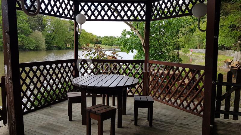 Kiosque en bois avec table et bancs, offrant une vue sur un étang entouré d'arbres verdoyants.