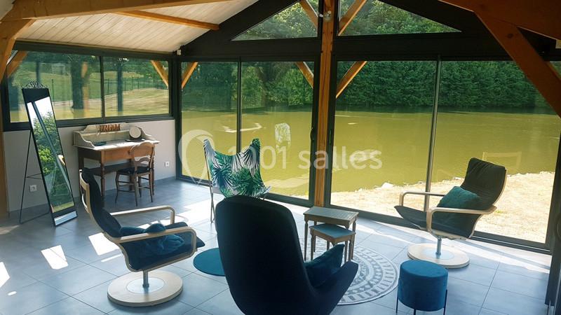 Véranda lumineuse avec vue sur un étang, aménagée avec fauteuils, table en bois et miroir sur pied.