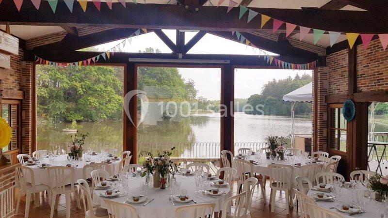 Salle décorée pour un événement, avec tables dressées et vue sur un lac entouré de verdure à travers de grandes baies…