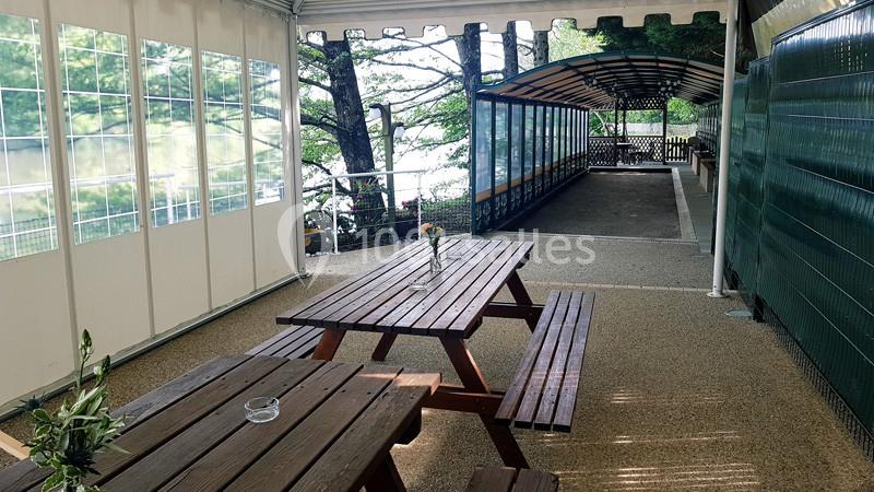 Tables en bois sous un abri ouvert avec vue sur des arbres et un chemin couvert en arrière-plan.