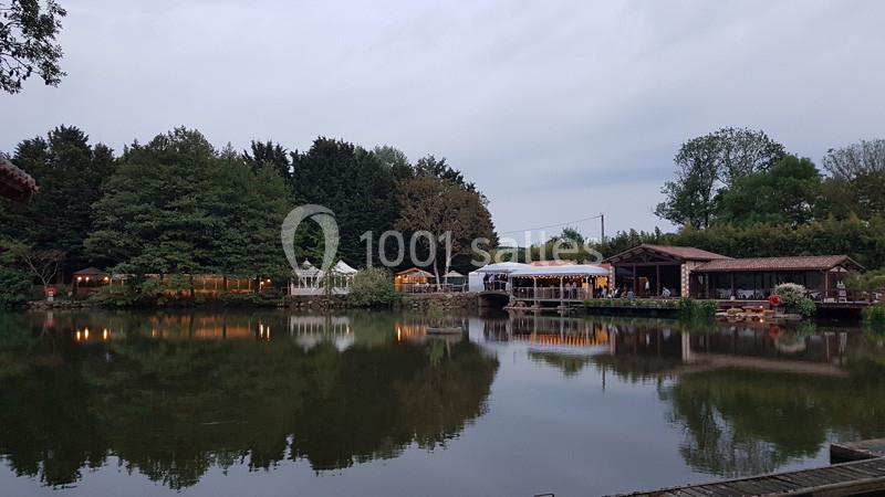 Étang entouré d'arbres avec des bâtiments en bois et des tentes éclairées se reflétant dans l'eau au crépuscule.