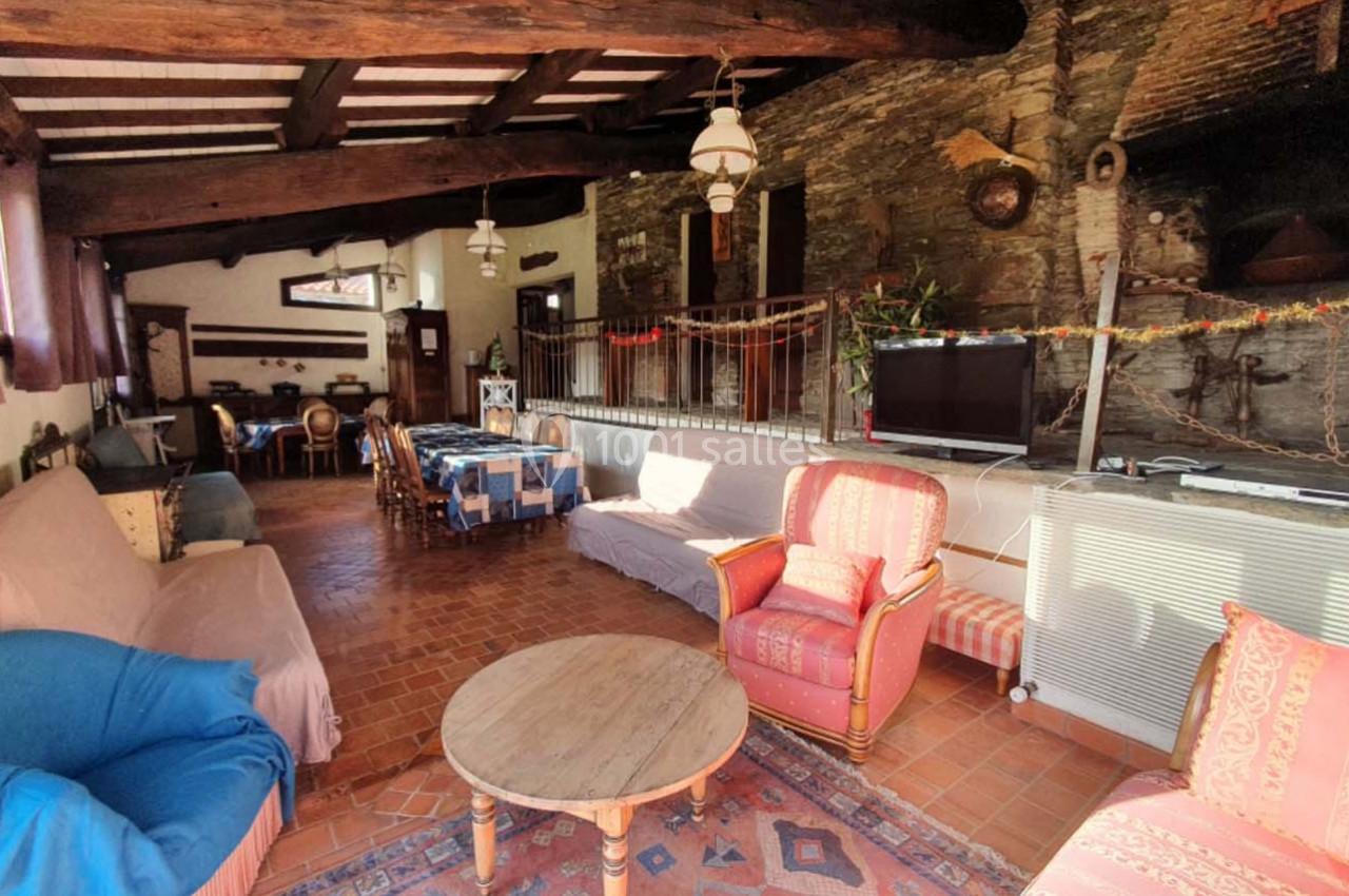 Salon rustique avec murs en pierre, poutres apparentes, canapés, fauteuils, table basse et coin repas en arrière-plan.