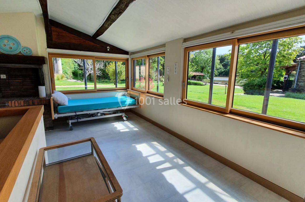 Véranda lumineuse avec grandes baies vitrées donnant sur un jardin verdoyant, mobilier simple et cheminée en brique.