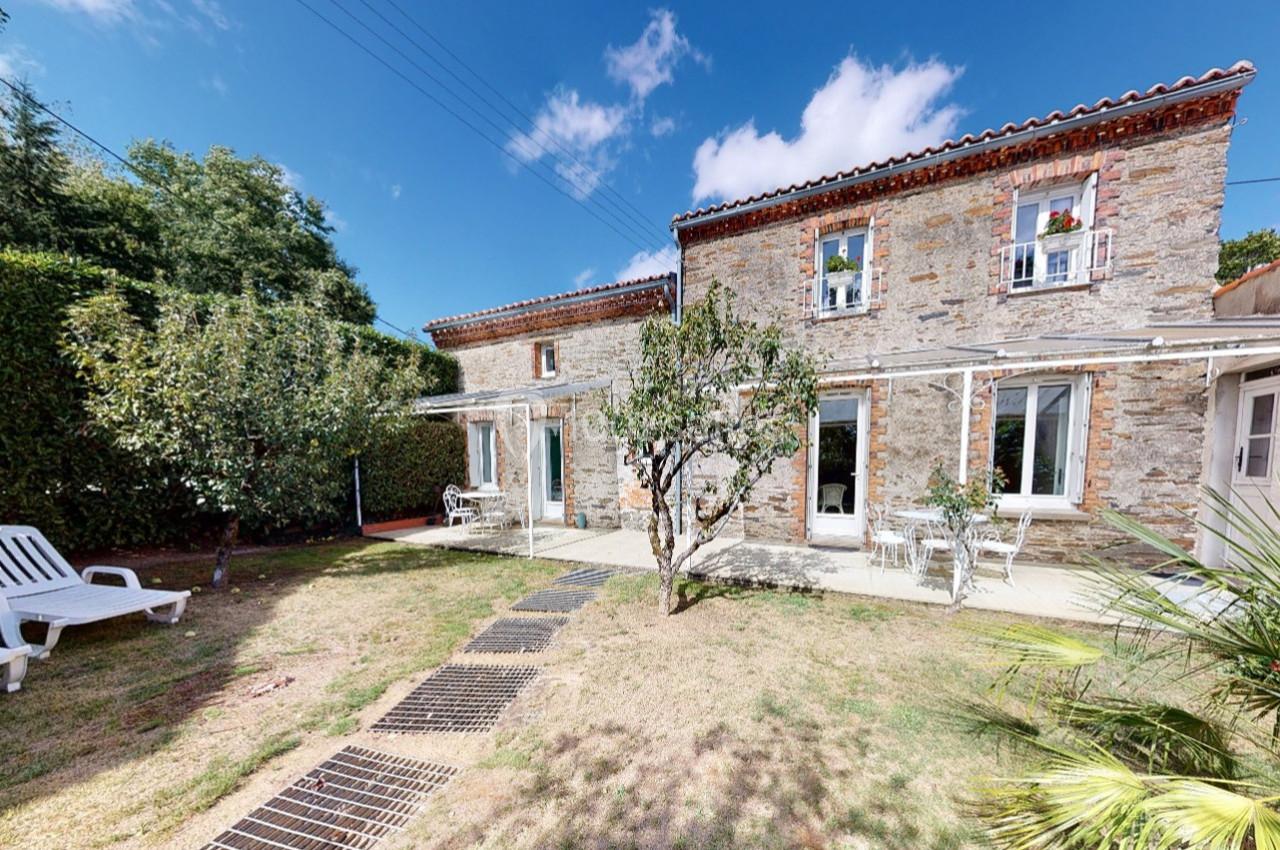 Façade en pierre d'une maison avec jardin, terrasse, chaises longues et arbres sous un ciel bleu.