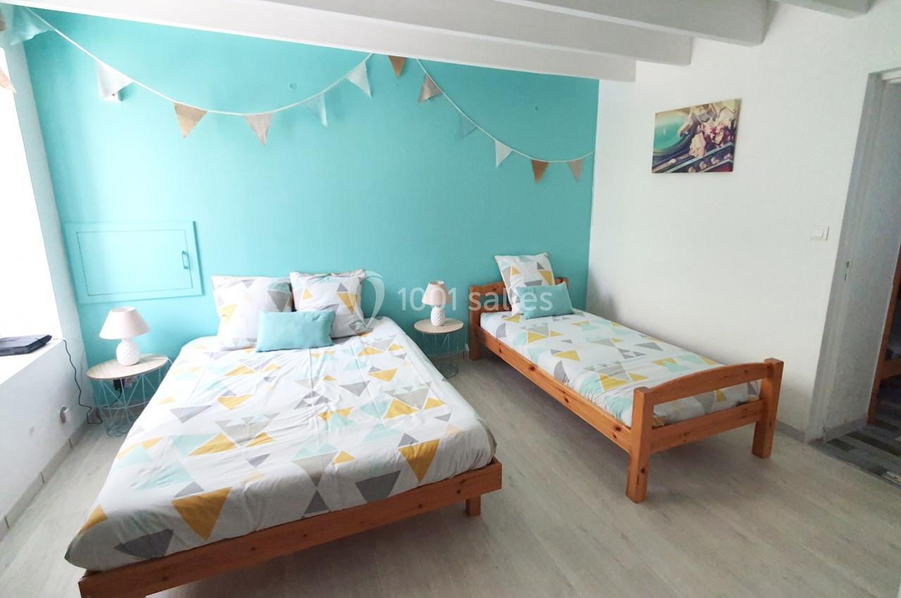 Chambre lumineuse avec un lit double, un lit simple, murs turquoise, guirlande décorative et parquet clair.
