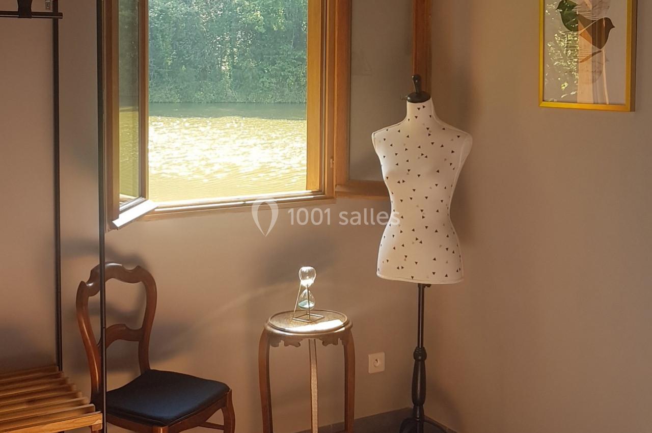 Mannequin de couture, chaise en bois et sablier sur une table près d'une fenêtre ouverte donnant sur un paysage verdoyant.