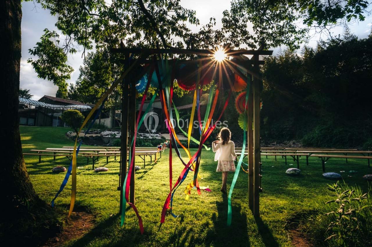 Une enfant marche sous une arche décorée de rubans colorés dans un jardin ensoleillé avec des bancs en arrière-plan.