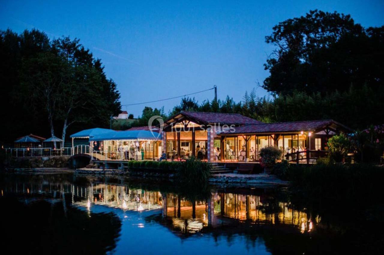 Bâtiment éclairé avec de grandes baies vitrées, au bord d'un étang, entouré de végétation, en soirée.