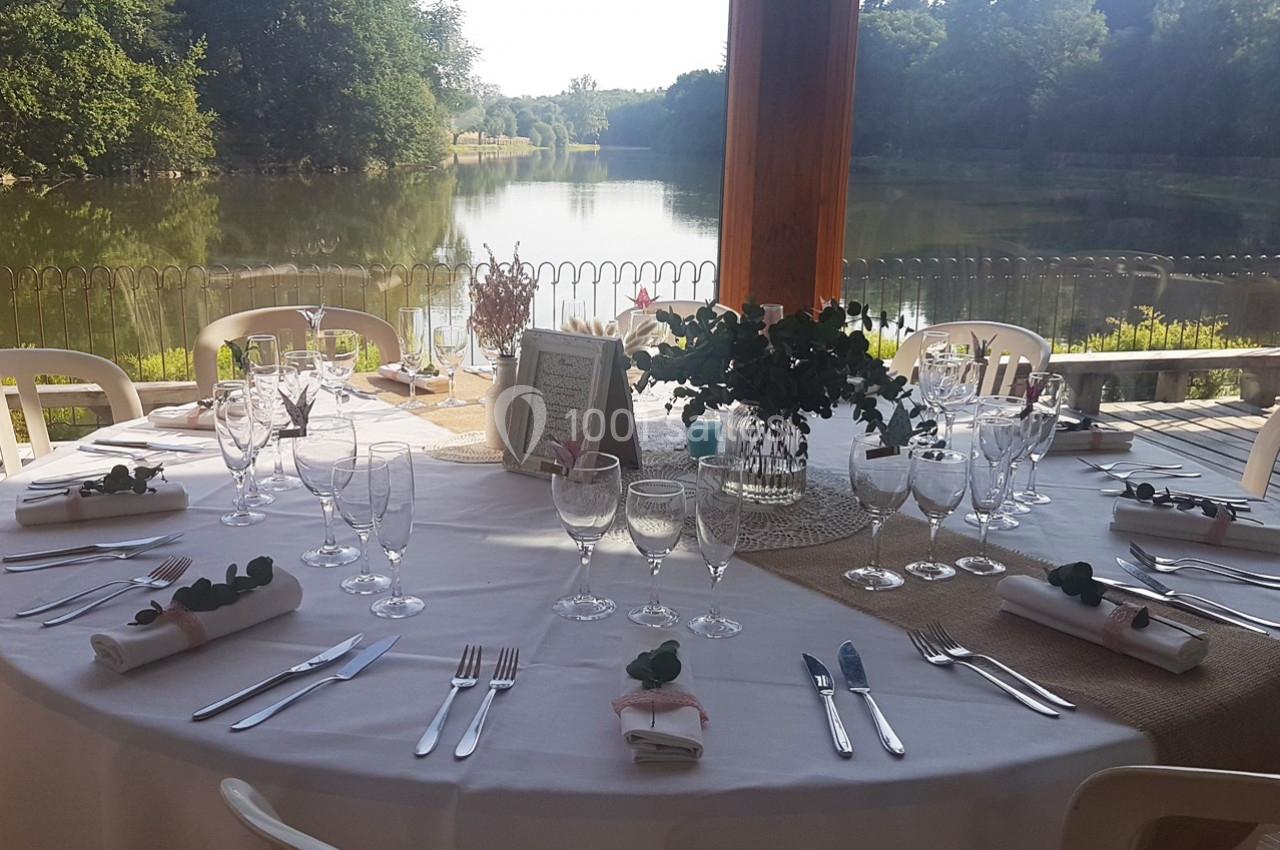 Table ronde élégamment dressée avec vaisselle et décoration florale, située en terrasse avec vue sur un lac entouré de…