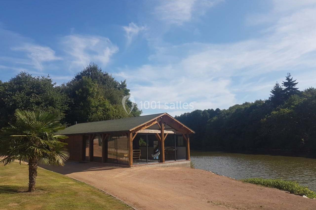 Chalet en bois avec toit vert, situé au bord d'un étang entouré d'arbres sous un ciel bleu.