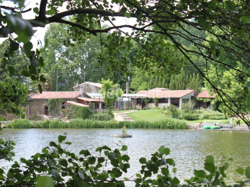 Vue d'un bâtiment en pierre avec toits en tuiles, entouré de verdure, au bord d'un étang avec une petite fontaine.