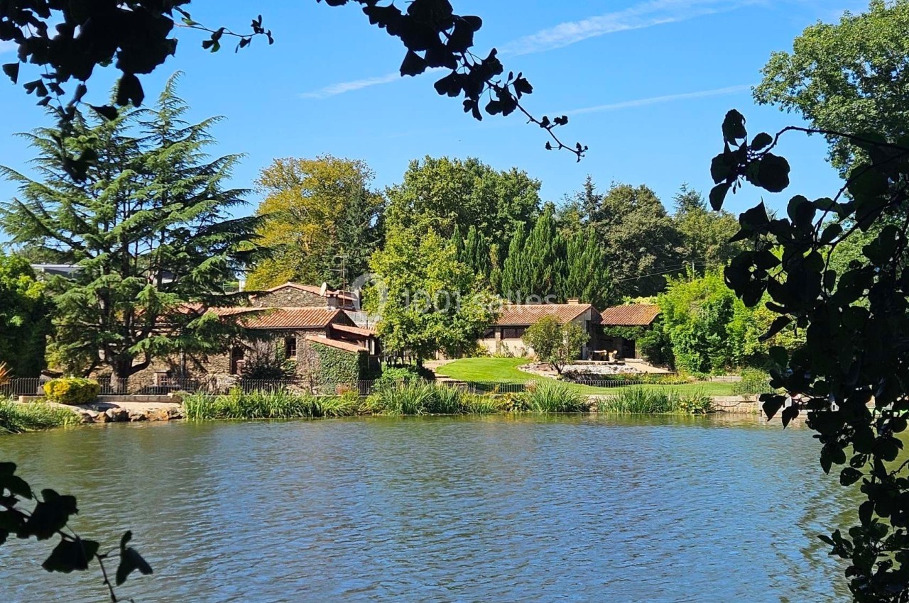 Vue d'un étang entouré de végétation, avec des bâtiments en pierre et des toits en tuiles au second plan.