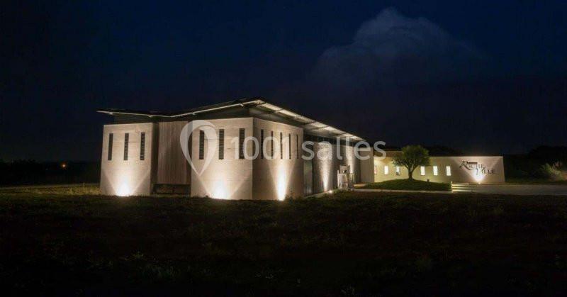 Bâtiment moderne éclairé la nuit, entouré d'un espace extérieur sombre avec un ciel nuageux en arrière-plan.
