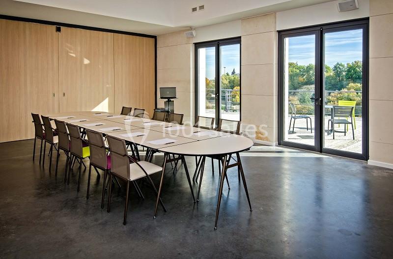 Salle de réunion lumineuse avec une grande table ovale, des chaises, un écran et des baies vitrées donnant sur une terrasse.