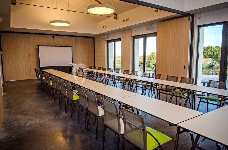 Salle de réunion lumineuse avec de longues tables alignées, des chaises et un écran de projection à l'avant.