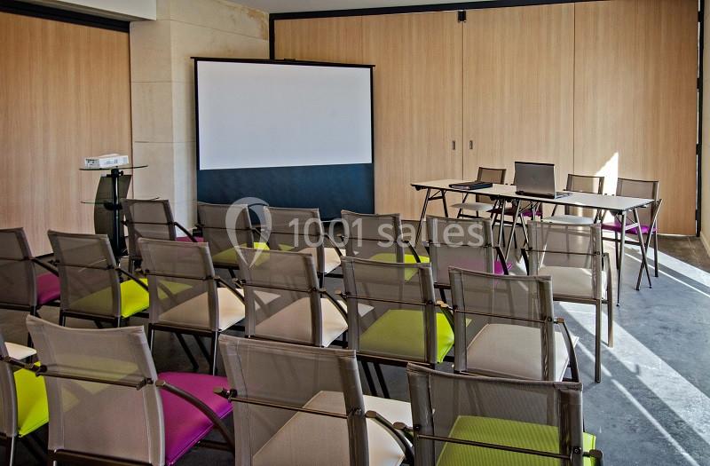 Salle de réunion avec des chaises colorées alignées face à un écran de projection et une table.
