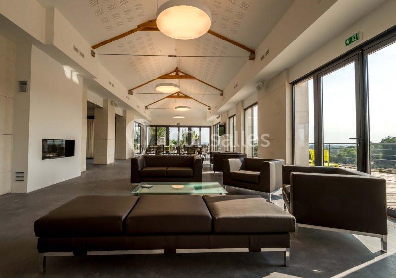 Salle lumineuse avec canapés en cuir, table basse en verre, grandes baies vitrées et vue sur un paysage extérieur.