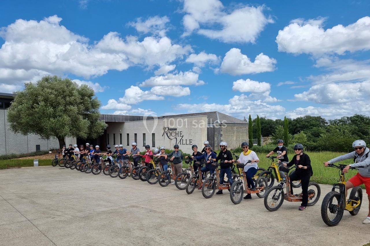 Un groupe de personnes alignées sur des trottinettes tout-terrain devant un bâtiment sous un ciel partiellement nuageux.