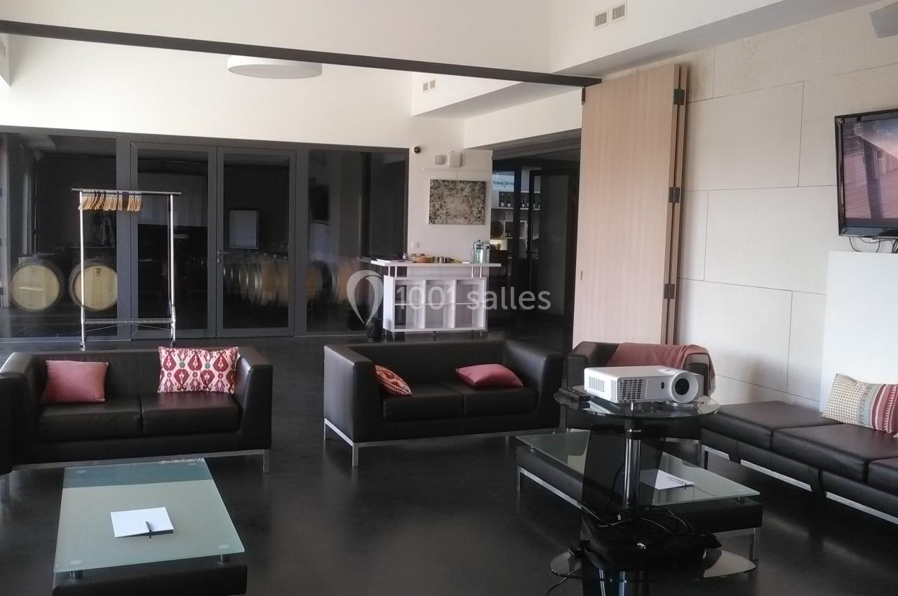 Salle moderne avec canapés noirs, tables basses en verre, projecteur, et bar en arrière-plan.