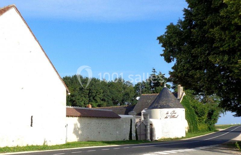 Mur blanc avec portail en fer forgé, bâtiment à toit conique et arbre bordant une route sous un ciel dégagé.