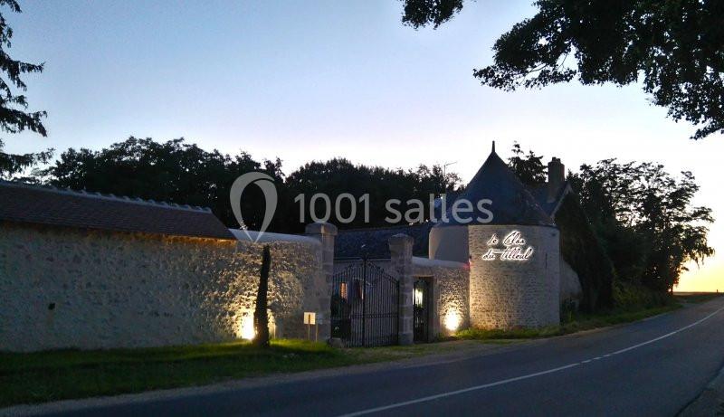 Façade en pierre éclairée d'un bâtiment avec une tour et une enseigne lumineuse, bordée par une route au crépuscule.
