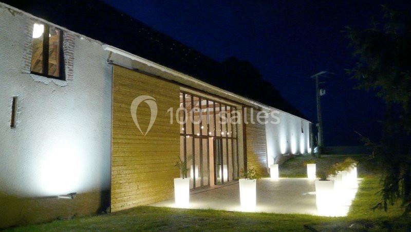 Façade d'une grange rénovée éclairée par des luminaires extérieurs, avec des pots lumineux alignés sur une terrasse.