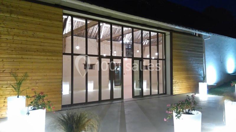 Façade vitrée d'un bâtiment moderne en bois, éclairée par des lumières extérieures et entourée de plantes en pots.