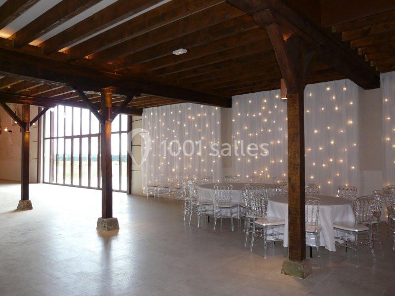 Salle avec poutres en bois, tables rondes et chaises transparentes, éclairée par des guirlandes lumineuses et une grande…