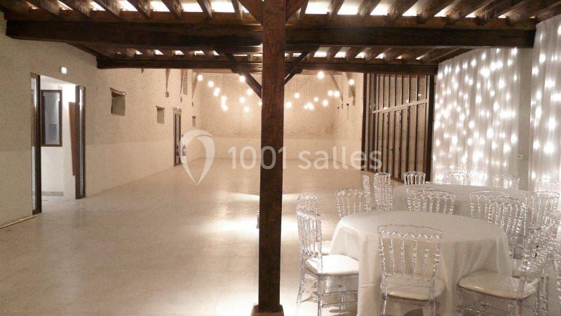 Salle spacieuse avec poutres en bois, murs blancs, éclairage suspendu et tables rondes avec chaises transparentes.