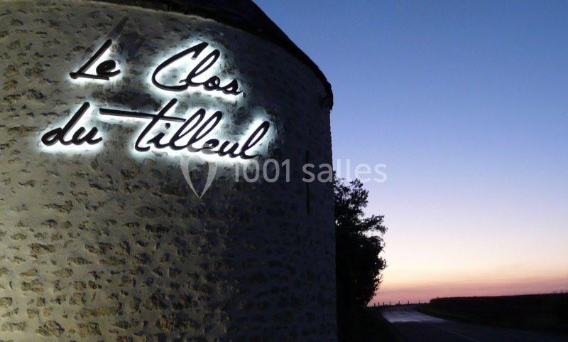 Mur en pierre avec une enseigne lumineuse ’Le Clos du Tilleul’, route et ciel au crépuscule en arrière-plan.