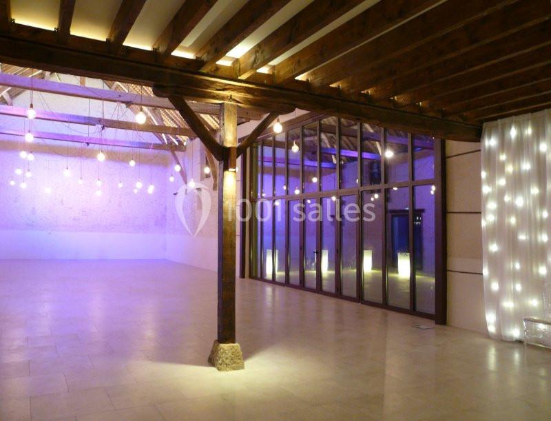 Salle spacieuse avec poutres apparentes, éclairage suspendu et murs vitrés donnant sur l'extérieur.