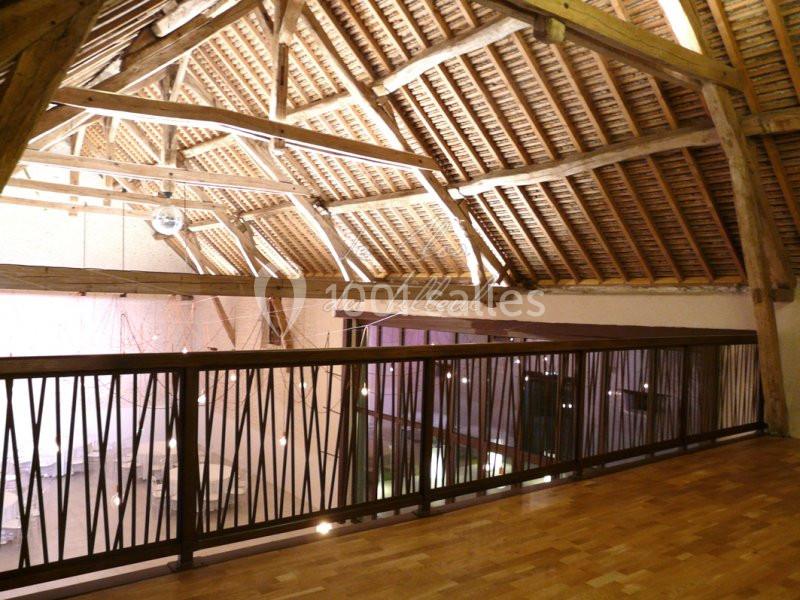 Charpente en bois apparente d'une salle avec mezzanine, parquet au sol et balustrade en métal.