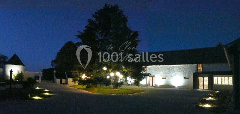 Cour éclairée d'un domaine avec bâtiments en pierre, un grand arbre central et un petit édifice en arrière-plan.