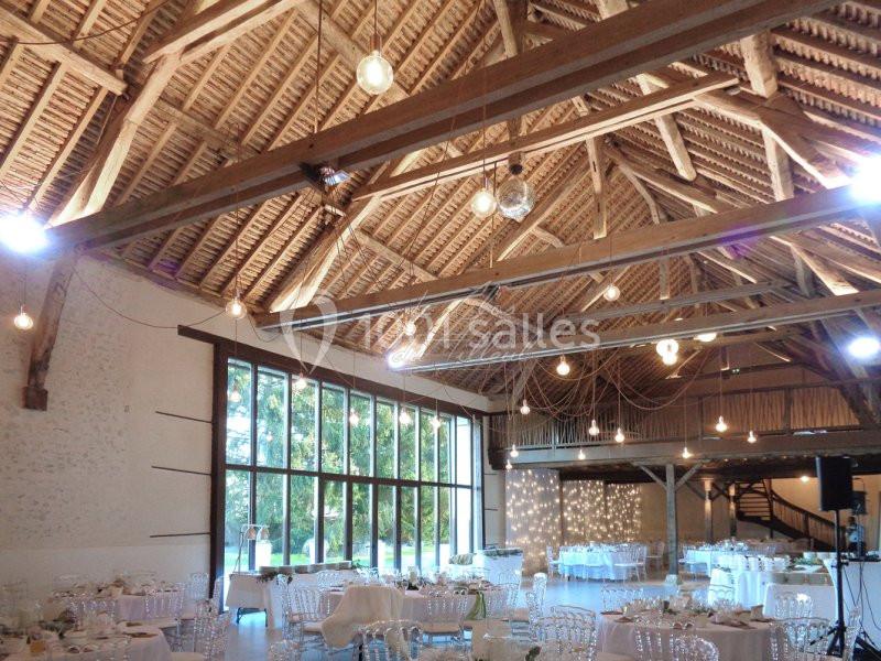 Salle de réception spacieuse avec charpente en bois, grandes baies vitrées et tables décorées pour un événement.