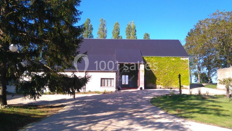 Bâtiment rural avec toit sombre, façade partiellement recouverte de lierre, entouré d'arbres et d'une allée gravillonnée.