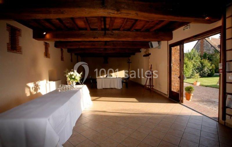 Salle lumineuse avec poutres apparentes, tables dressées et vue sur un jardin par une porte ouverte.