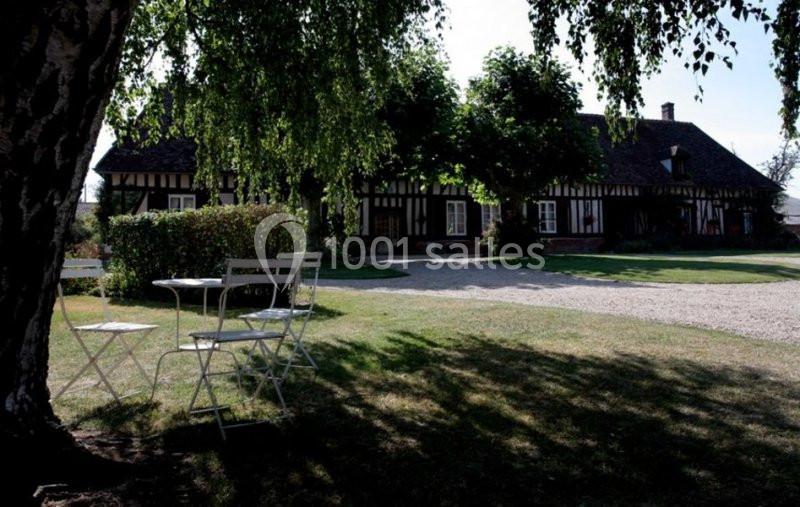 Maison à colombages entourée d'arbres, avec des tables et chaises blanches sur une pelouse ombragée.