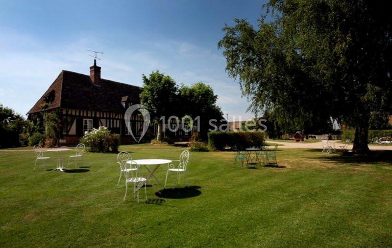 Jardin avec tables et chaises en fer forgé, devant une maison à colombages entourée d'arbres et de pelouse.