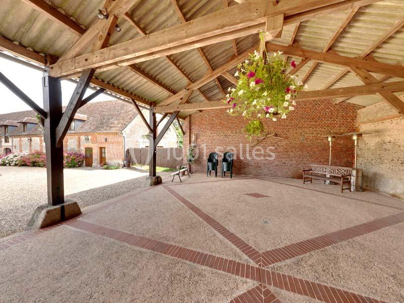 Structure en bois ouverte avec toit en tuiles, sol pavé, bancs, chaises et suspension florale, vue sur cour pavée.
