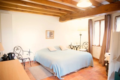 Chambre mansardée lumineuse avec lit simple, poutres apparentes, fenêtre, table de chevet et étagère en bois.