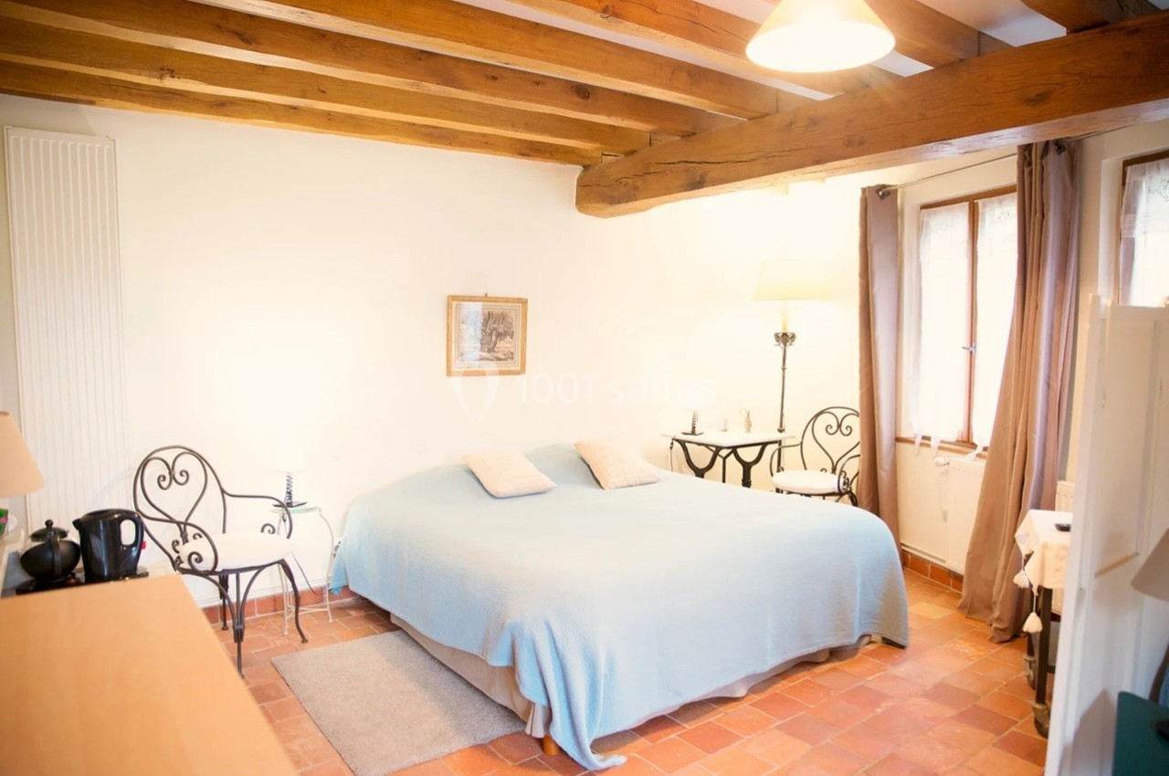 Chambre lumineuse avec lit double, poutres apparentes, sol en terre cuite, mobilier en fer forgé et fenêtre avec rideaux.