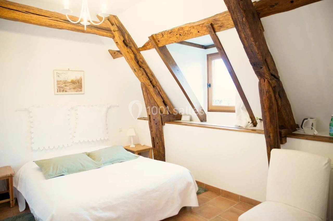 Chambre lumineuse avec poutres apparentes, lit double, murs blancs, fenêtre et décoration minimaliste.