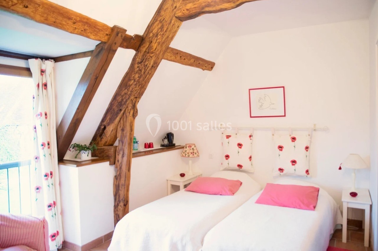 Chambre lumineuse avec deux lits simples, poutres apparentes, rideaux fleuris et décoration sobre.