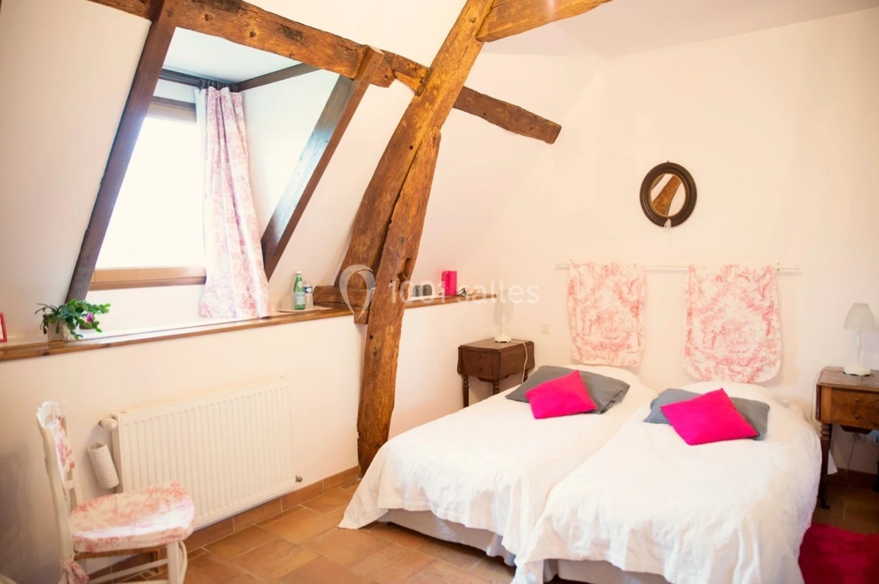 Chambre lumineuse avec poutres apparentes, deux lits simples, murs blancs et décoration rose.