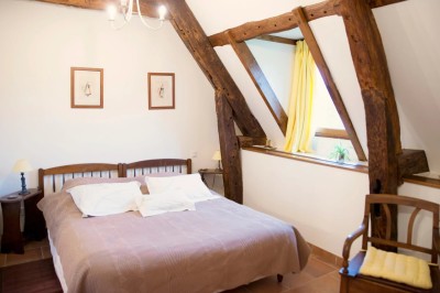 Chambre mansardée lumineuse avec lit simple, poutres apparentes, fenêtre, table de chevet et étagère en bois.