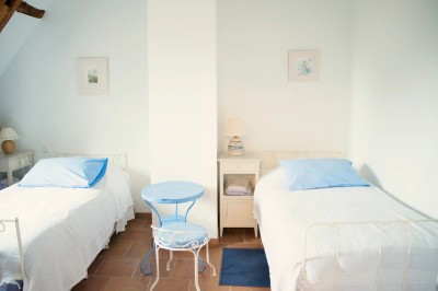 Chambre mansardée lumineuse avec lit simple, poutres apparentes, fenêtre, table de chevet et étagère en bois.