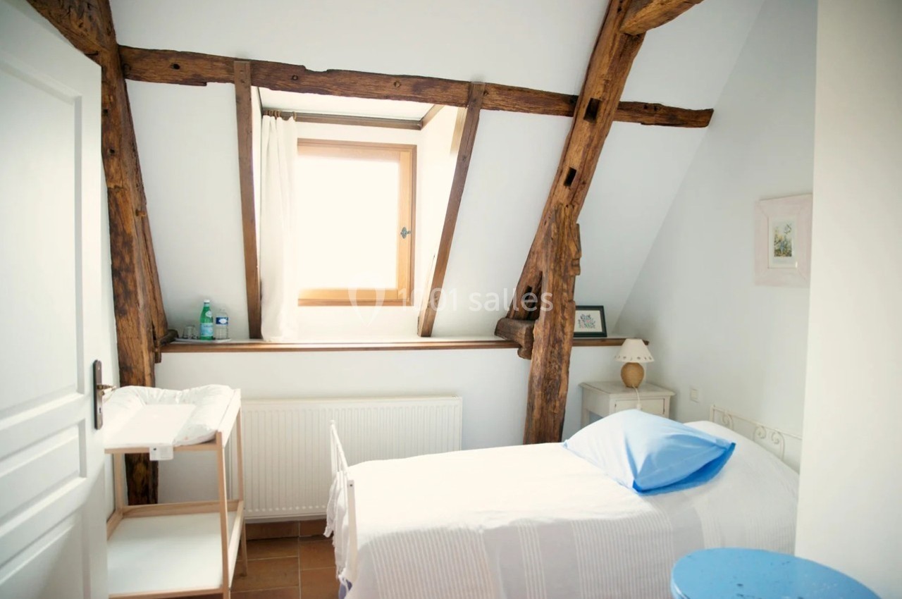 Chambre mansardée lumineuse avec lit simple, poutres apparentes, fenêtre, table de chevet et étagère en bois.