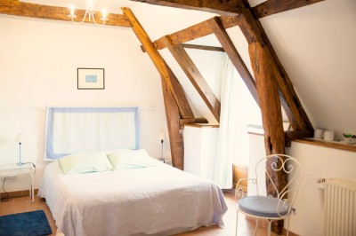 Chambre mansardée lumineuse avec lit simple, poutres apparentes, fenêtre, table de chevet et étagère en bois.