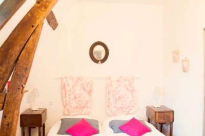 Chambre mansardée lumineuse avec lit simple, poutres apparentes, fenêtre, table de chevet et étagère en bois.