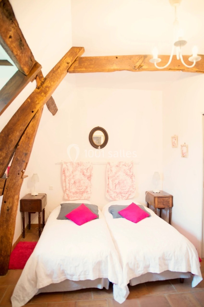 Chambre lumineuse avec poutres apparentes, deux lits simples côte à côte, coussins roses et décoration sobre.